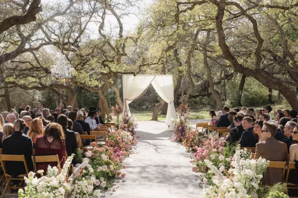 Ceremony aisle design features a flower-lined aisle under oak trees, with a chandelier shimmering above a stone path.