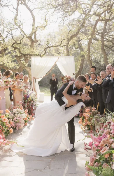 Ceremony kiss moment with a wedding dip kiss beneath a draped fabric arch, sunlight filtering through garden trees along a stone path.