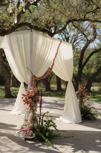 Draped wedding arch for outdoor ceremony decor, with fabric draping and hanging crystals, set beneath shady trees.