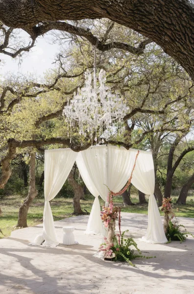 Ceremony arch with a crystal chandelier beneath an oak tree canopy on a stone patio, open sky peeking through.