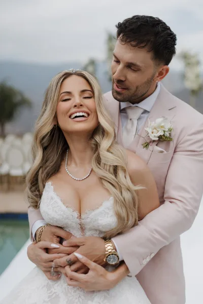 Couple portrait capturing the bride and groom embrace, her lace bodice catching light, set among palm trees with mountains and a blue sky.