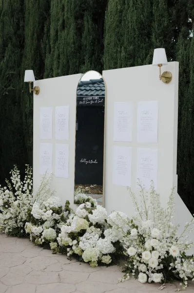 Wedding seating chart presented as a mirror seating chart, flanked by floral arrangements on a stone patio by garden trees.