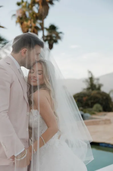 Couple portrait, an under the veil photo of the bride and groom embracing beneath a flowing bridal veil by the pool with palm trees and mountains.
