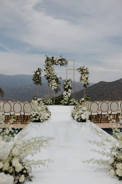 Ceremony setup with a modern wedding altar framed by a gold geometric arch, set outdoors with mountains under a clear sky.