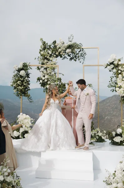 Ceremony moment during the wedding pronouncement as the couple raise hands beneath a gold frame arch, with mountains and sky beyond.