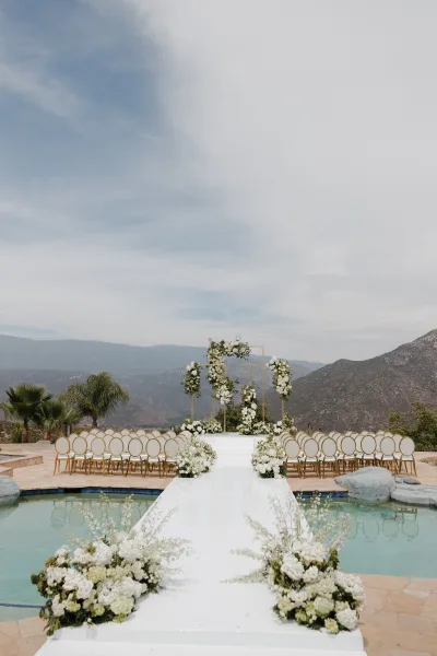 Ceremony setup showcasing outdoor ceremony decor on a poolside stone patio, rows of gold chairs facing mountains and palms.