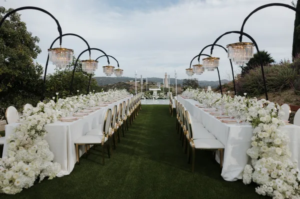 Reception tablescape for outdoor reception decor, set beneath chandeliers on a manicured lawn with trees and rolling hills under open sky.