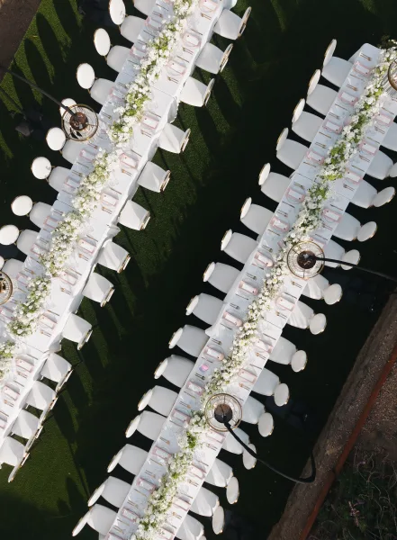 Reception tablescape on long banquet tables under pendant lights, set on a grass lawn beside a pathway.