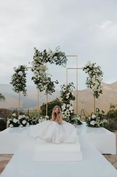 Bridal portrait of a seated bride beneath a white floral arch, set against distant mountains and trees under a clear sky.