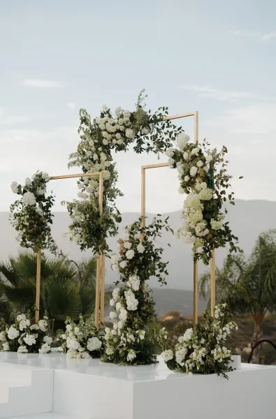 Wedding arch serving as a geometric ceremony backdrop, trimmed with white florals, stands against mountains and palm trees under a blue sky.