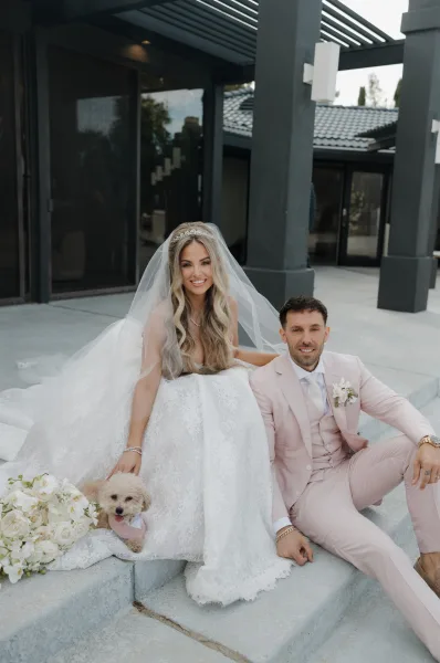 Couple portrait of a bride and groom with dog on concrete steps, the pup in a bow tie, framed by modern architecture and glass doors.