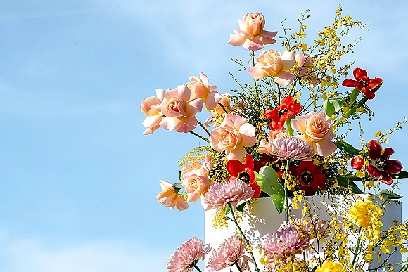 Wedding cake flowers and cake floral topper with peach roses, red tulips, pink chrysanthemums, and yellow orchids on a white cake, blue sky backdrop