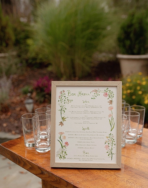 Wedding bar menu sign in a frame with floral illustrations on a wooden table beside glasses, set against lush garden greenery and blooms