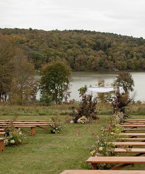 Outdoor ceremony setup with lakeside wedding ceremony arch draped in fabric, wood benches, aisle florals, and pumpkins on a grassy lawn by the lake