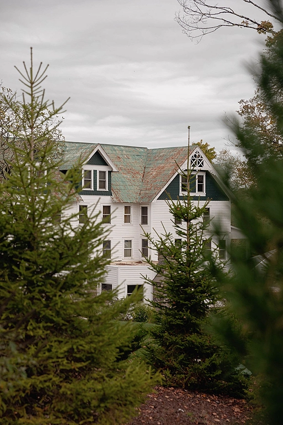 Wedding venue exterior with gable roof and dormer windows, set along a gravel driveway with evergreen trees under a cloudy sky