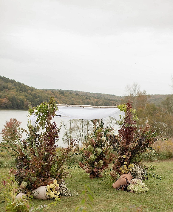 Wedding ceremony backdrop with floral wedding arch of birch poles and white draping, greenery and ground florals by a lake and hills