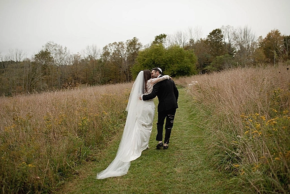 Wedding couple portrait of bride and groom walking away down a dirt path, her long veil trailing over meadow grass and wildflowers