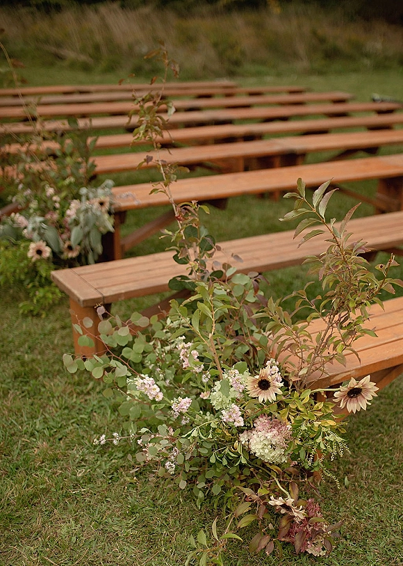 Ceremony seating with wood bench ceremony seating on a meadow lawn, with a floral ground arrangement of eucalyptus, sunflowers, and hydrangea