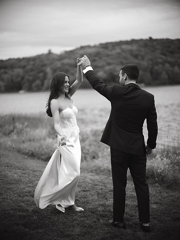 Couple portrait of newlyweds twirling by a lakeside shoreline, bride in a strapless gown holding her hem as groom leads under cloudy hills