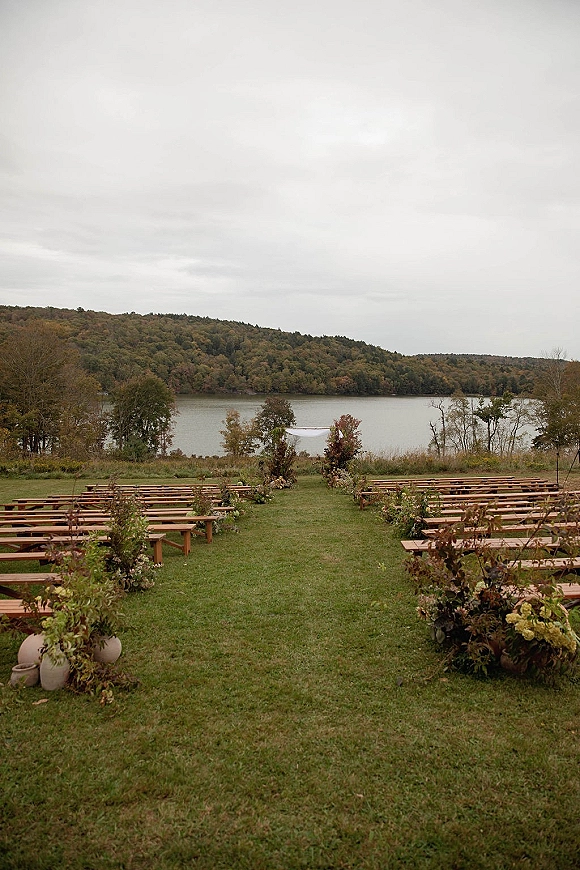 Outdoor ceremony setup with wood benches and aisle florals leading to a white draped arch on a lakeside lawn under overcast hills