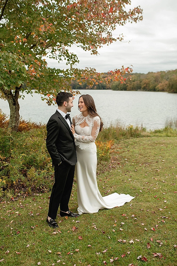 Couple portrait of bride and groom laughing by a lakeside lawn, her long sleeve lace dress train beside his tuxedo under fall foliage
