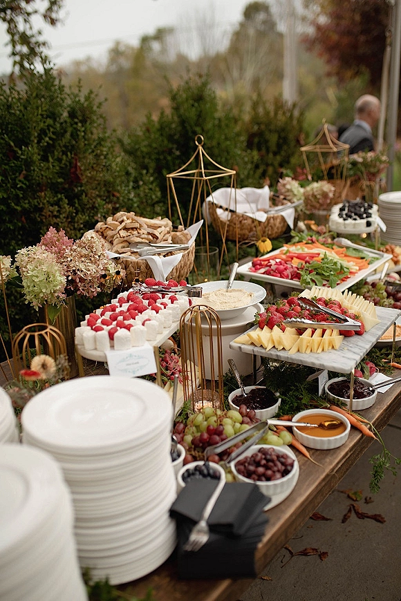 Wedding dessert table with a wedding grazing table spread of cheeses, fruit, crackers and marshmallows on marble boards, set by garden hedges