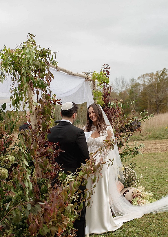 Wedding vows as groom in black suit and kippah reads into a handheld microphone under a white-draped outdoor chuppah in a grassy field