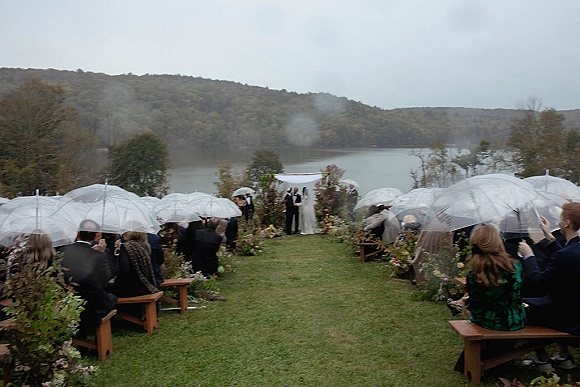 Outdoor wedding ceremony in a rainy wedding ceremony with guests under clear umbrellas, wooden benches, and a floral arch by the lake