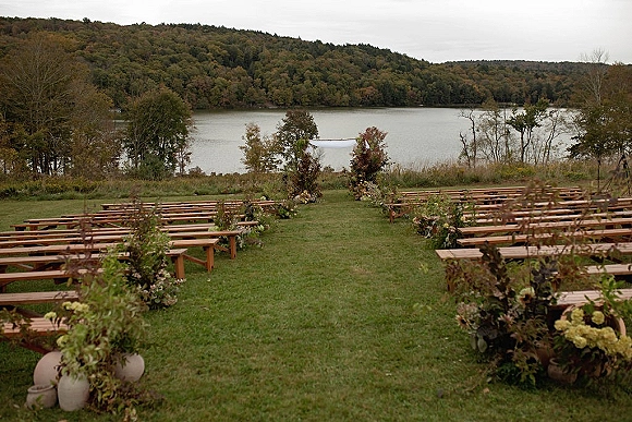 Ceremony setup for an outdoor wedding ceremony with wood benches lining a white aisle runner, florals and greenery facing a lakeside lawn backdrop