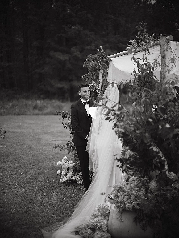 Ceremony moment as bride and groom at altar under a draped fabric arch with greenery garland on a lawn framed by trees