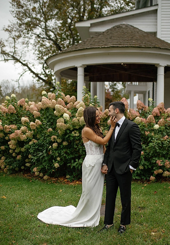 Couple portrait of bride and groom holding hands, bride in lace gown with train and ring beside hydrangea bushes by a gazebo