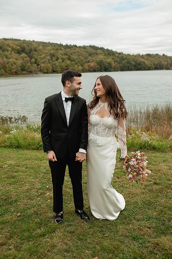 Couple portrait of bride and groom holding hands, walking by a lake on grassy shore under overcast sky, bouquet in hand