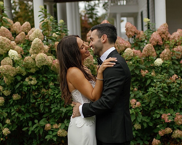 Couple portrait of bride and groom embrace, forehead to forehead, in lace dress and black tuxedo before pink hydrangeas and porch columns