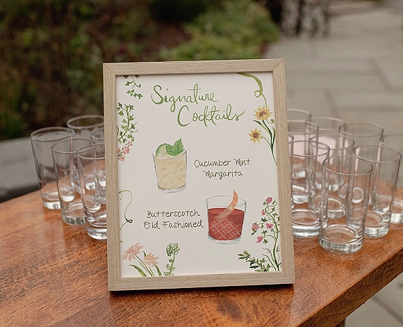 Signature cocktail sign in a wood frame with illustrated wedding bar menu sign, watercolor florals and glasses on a wooden table outdoors