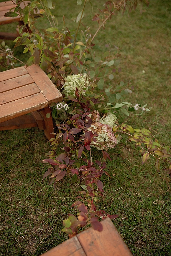 Aisle floral decor with ceremony aisle flowers, hydrangea blooms and greenery garland spilling beside a wood bench on a grass lawn