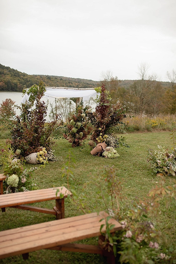 Ceremony arch decor with white drapery and floral greenery on a wood arch, framed by ceramic vases on a lakeside lawn with hills