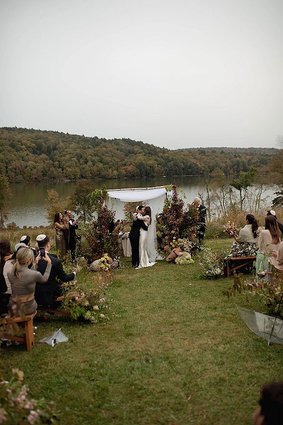 Ceremony kiss moment as bride and groom kiss under a white-draped wedding arch with florals on a lakefront lawn with benches