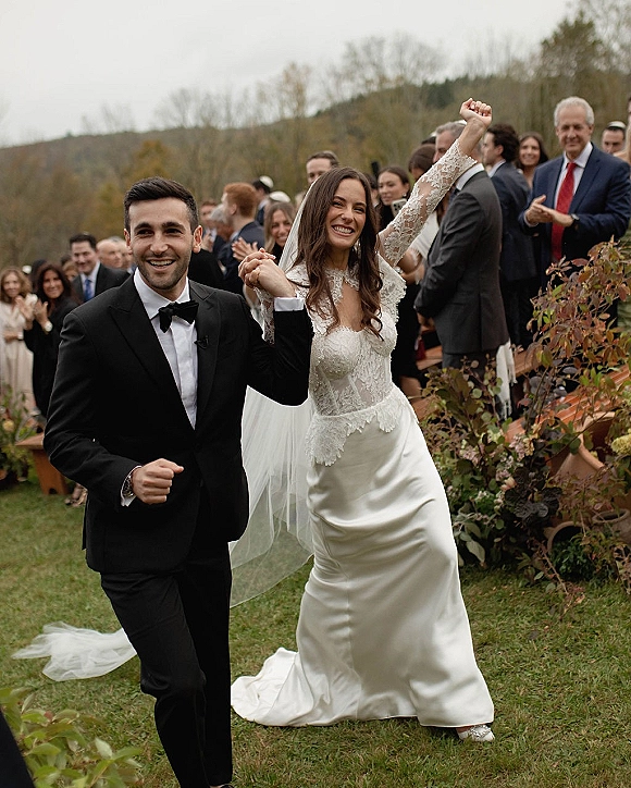 Wedding recessional as bride and groom walk the aisle hand in hand, bride in lace long sleeves and veil, guests cheer on wooden benches outdoors