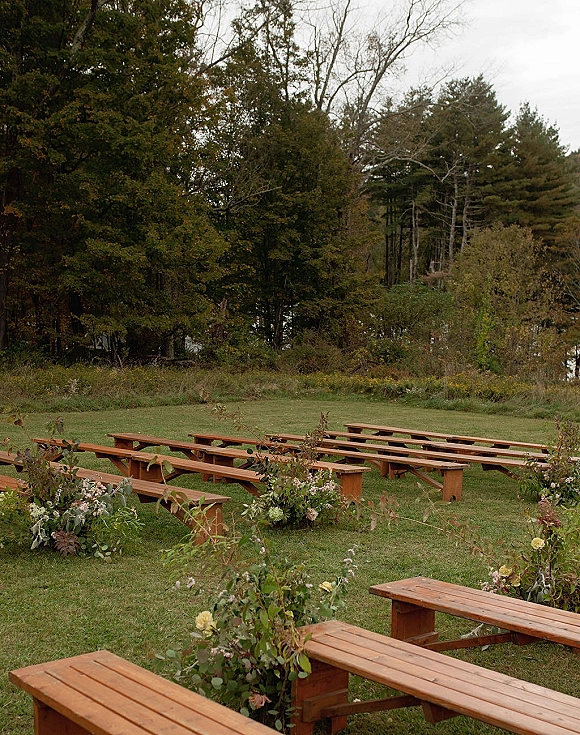 Ceremony seating with wood benches lined by greenery and floral arrangements on a grassy lakeside lawn under a cloudy sky