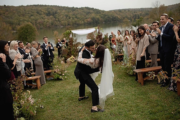 Wedding kiss as newlyweds dip in the aisle, bride’s veil flowing, guests cheering by rustic benches at a lakeside ceremony under overcast skies
