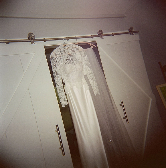 Wedding dress with lace sleeves hangs on a hanger, satin skirt draped beneath a bridal veil against wardrobe doors in a bedroom wall