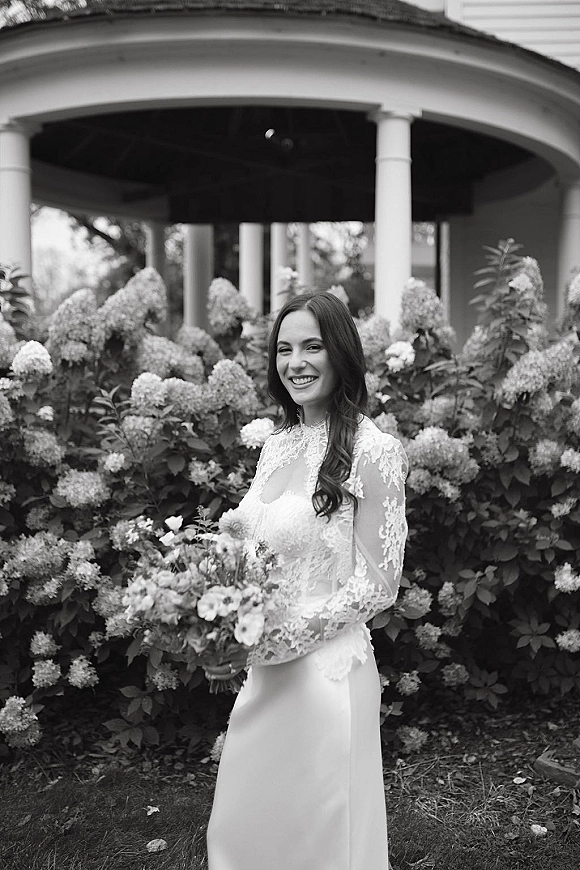 Bridal portrait of a smiling bride holding bouquet in a lace long sleeve, high neck gown by hydrangea bushes near a gazebo column