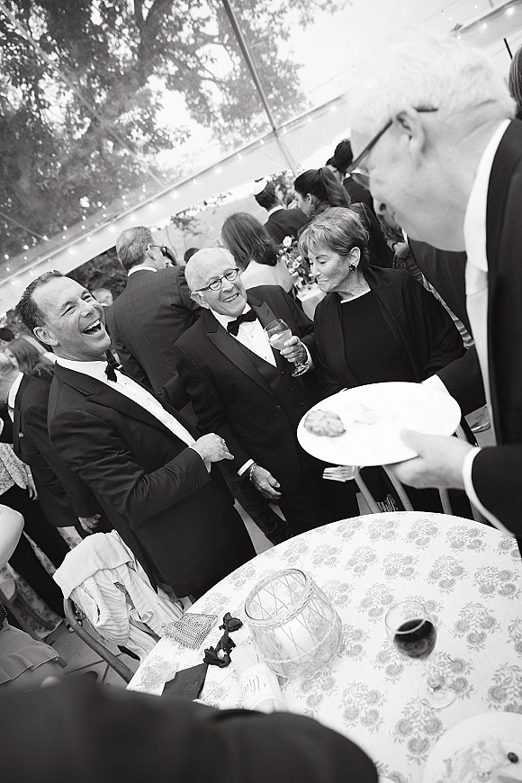 Wedding reception guests at cocktail hour laughing and chatting in tuxedos, holding wine glasses under string lights in a clear tent
