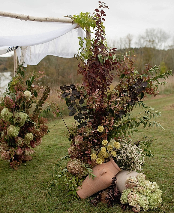 Wedding ceremony arch with white drapery and lush florals in ceramic vases, set on a grassy lawn by water under overcast sky