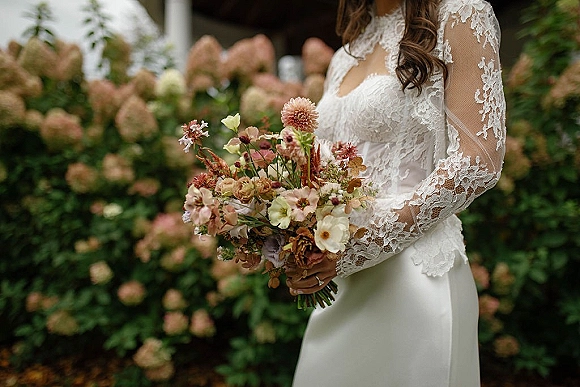 Bridal bouquet of airy wildflowers and greenery wrapped with brown ribbon, held against a long-sleeve lace dress on a garden walkway