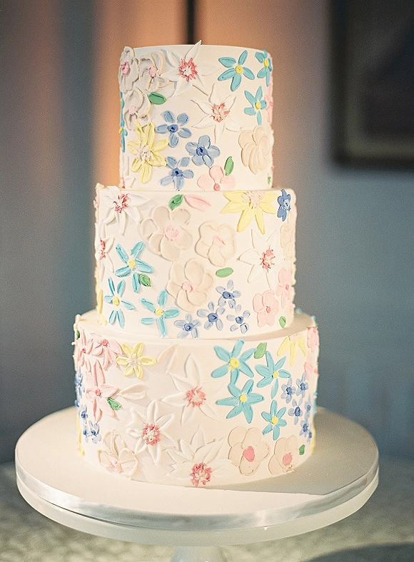 Wedding cake with three tier wedding cake design, textured buttercream frosting and pastel piped flowers on a stand in soft natural light indoors
