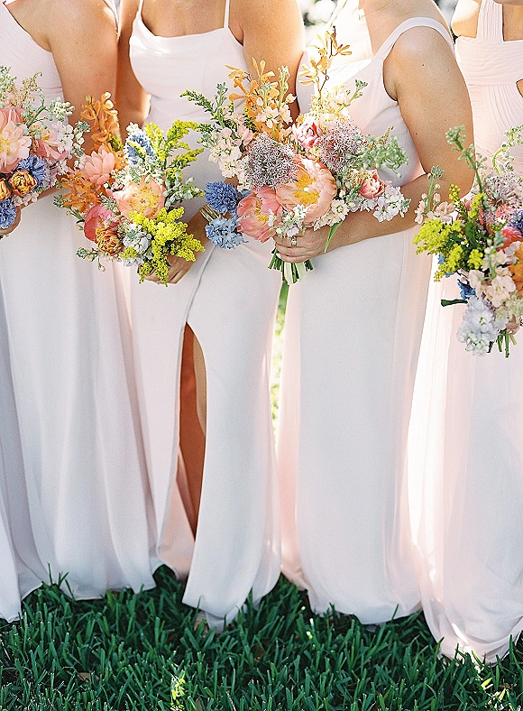 Bridesmaid bouquets in a row, colorful bridesmaid bouquets with wildflowers held by women in spaghetti-strap dresses on green grass