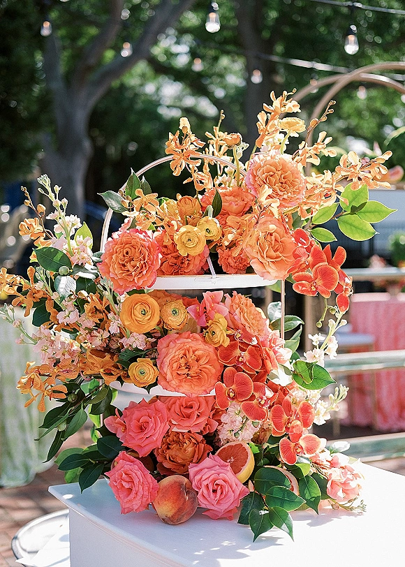 Wedding floral centerpiece with orange wedding flowers on a tiered white stand, roses, orchids, greenery, and citrus slice on a patio under string lights