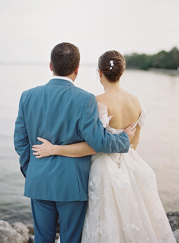 Couple portrait of bride and groom from behind, hugging on a rocky beach shoreline, her lace off-shoulder gown and floral hair pins visible