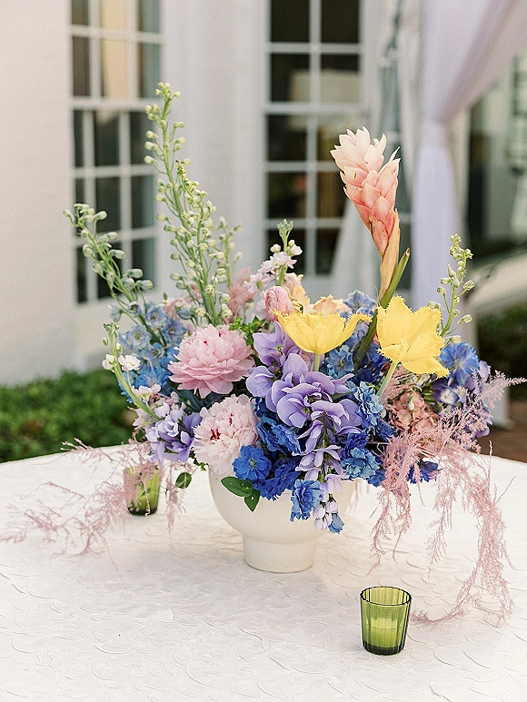 Wedding centerpiece with a pastel floral arrangement in a white ceramic vase, accented by a green glass votive on a white tablecloth